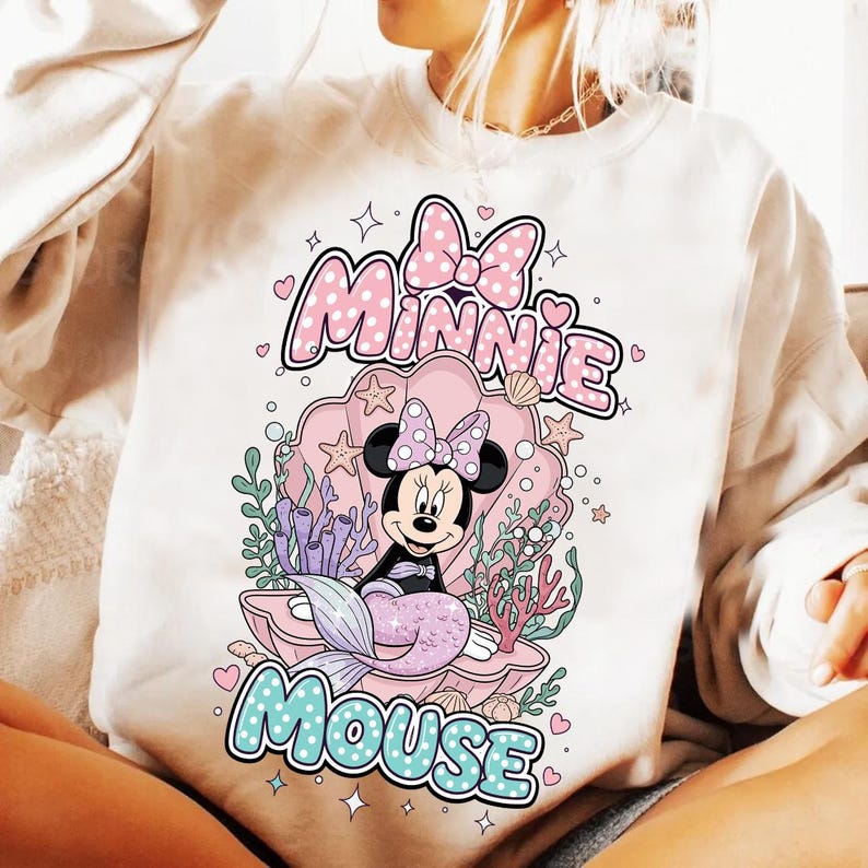 May include: Cream-colored sweatshirt featuring a Minnie Mouse mermaid graphic. The design includes the words "Minnie Mouse" in a bubble font, a pink seashell, and aquatic elements. Minnie is depicted with a purple tail and a polka-dot bow.