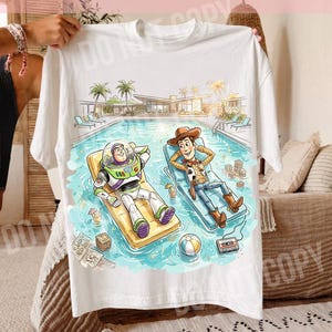 May include: White t-shirt featuring a colorful illustration of Buzz Lightyear and Woody relaxing in a pool. The design includes a pool scene with palm trees, a building, and various accessories. The text "ORIGINAL DESIGNER CALIGRAPHICDESIGN" is at the top.