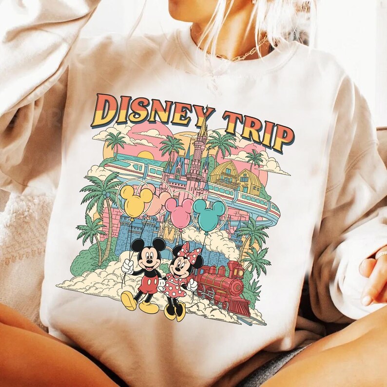May include: Cream-colored sweatshirt with a colorful graphic that says "DISNEY TRIP." The graphic features Mickey and Minnie Mouse, a castle, train, balloons, and palm trees. The design has a vintage, retro style.