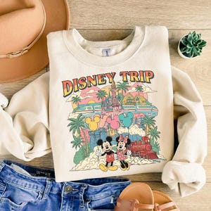 May include: Cream-colored sweatshirt with a colorful graphic that says "DISNEY TRIP." The graphic features Mickey and Minnie Mouse, a castle, palm trees, balloons, and a train. The sweatshirt is paired with blue denim shorts, a hat, and sandals.