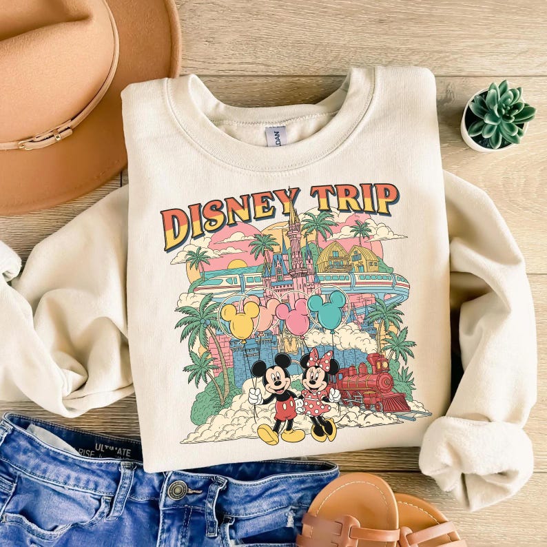 May include: Cream-colored sweatshirt with a colorful graphic that says "DISNEY TRIP." The graphic features Mickey and Minnie Mouse, a castle, palm trees, balloons, and a train. The sweatshirt is paired with blue denim shorts, a hat, and sandals.