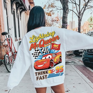 May include: White sweatshirt featuring a graphic of Lightning McQueen from the Cars movie. The design includes the text "Lightning McQueen", "95", and "Think Fast", along with checkered flags and a red race car.