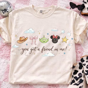May include: Cream-colored t-shirt featuring a whimsical design with a cowboy hat, a pink bow, a green alien, a Minnie Mouse head, and a yellow star. The text "you got a friend in me!" is written in a script font.