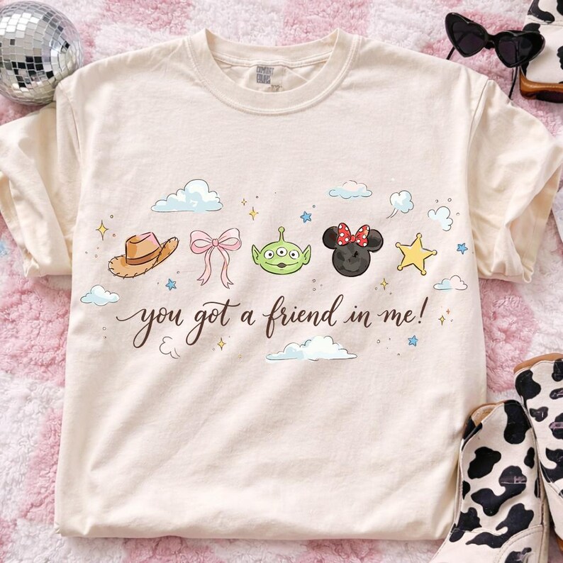 May include: Cream-colored t-shirt featuring a whimsical design with a cowboy hat, a pink bow, a green alien, a Minnie Mouse head, and a yellow star. The text "you got a friend in me!" is written in a script font.