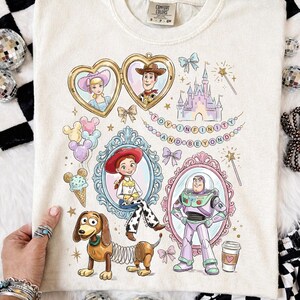 Może przedstawiać: Kremowa koszulka z kolorowymi ilustracjami postaci z Toy Story, w tym Chudego, Buzz'a Astrala i Jessie, w ozdobnych ramkach. Fraza "TO INFINITY AND BEYOND" jest wyświetlana u g&oacute;ry. Koszulka ma metkę Comfort Colors.