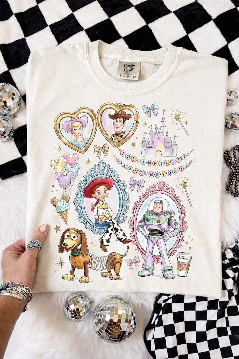 Może przedstawiać: Kremowa koszulka z kolorowymi ilustracjami postaci z Toy Story, w tym Chudego, Buzz'a Astrala i Jessie, w ozdobnych ramkach. Fraza "TO INFINITY AND BEYOND" jest wyświetlana u g&oacute;ry. Koszulka ma metkę Comfort Colors.