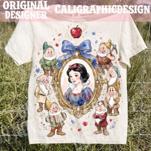 May include: A cream-colored short-sleeve t-shirt featuring a central graphic of a woman with black hair in a gold ornate frame topped with a blue bow. Seven small male figures in various poses surround the frame, with a red apple above the bow.