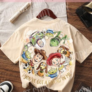 以下が含まれることがあります： アニメ映画「トイ・ストーリー」のキャラクターが描かれた、ベージュ色のTシャツ。ウッディ、バズ・ライトイヤー、ジェシーなどのキャラクターと「You've Got a Friend in Me」の文字がデザインされています。