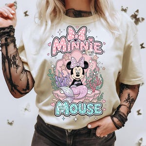 May include: Cream-colored t-shirt featuring a Minnie Mouse mermaid design. The graphic includes Minnie in a seashell, surrounded by coral and seaweed. The text "Minnie Mouse" is displayed above and below the image in pink and blue.