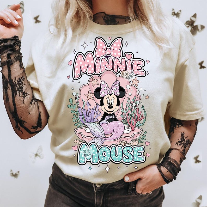 May include: Cream-colored t-shirt featuring a Minnie Mouse mermaid design. The graphic includes Minnie in a seashell, surrounded by coral and seaweed. The text "Minnie Mouse" is displayed above and below the image in pink and blue.