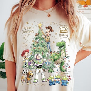 May include: Cream-colored t-shirt featuring a festive Toy Story holiday design. The graphic includes Woody, Buzz Lightyear, Rex, Jessie, and other characters decorating a Christmas tree. Text reads "Merry Christmas" and "Toy Story Holiday".