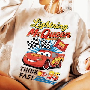 May include: White sweatshirt featuring a graphic of Lightning McQueen from the Cars movie. The design includes the text "Lightning McQueen", "Think Fast", and the number "95". The image also shows checkered flags and a red race car.
