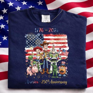 K&ouml;nnte beinhalten: Marineblaues Sweatshirt mit einer Grafik der amerikanischen Flagge, Feuerwerk und dem Text "1776-2026". Die Grafik enth&auml;lt Zeichentrickfiguren. Der Text "America - 250th Anniversary" ist ebenfalls vorhanden.