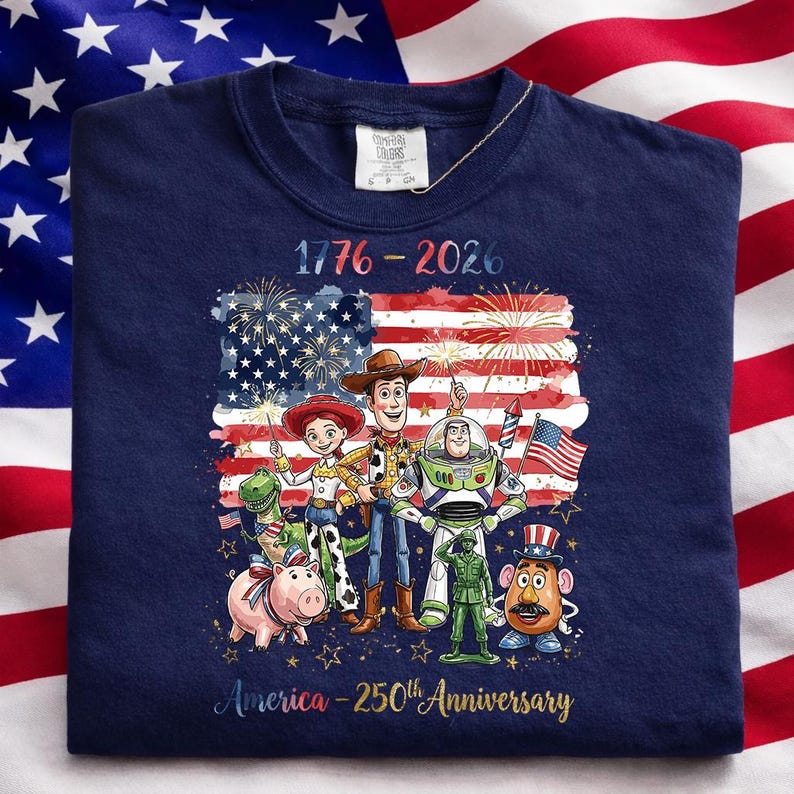 K&ouml;nnte beinhalten: Marineblaues Sweatshirt mit einer Grafik der amerikanischen Flagge, Feuerwerk und dem Text "1776-2026". Die Grafik enth&auml;lt Zeichentrickfiguren. Der Text "America - 250th Anniversary" ist ebenfalls vorhanden.