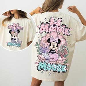 May include: Cream-colored t-shirt featuring a Minnie Mouse mermaid design. The graphic includes a pink bow, the words "Minnie Mouse", and a mermaid tail. The design is repeated on the front of the shirt.