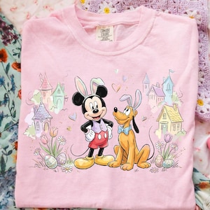 Può includere: T-shirt rosa Comfort Colors con Topolino e Pluto vestiti per Pasqua, con orecchie da coniglio. La grafica include castelli dai colori pastello, uova di Pasqua, fiori e cuori. La maglietta è su una superficie a motivi floreali.