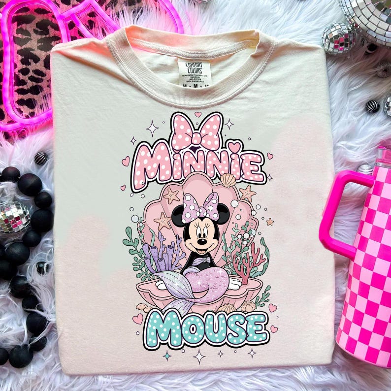May include: Cream-colored t-shirt featuring a graphic of Minnie Mouse as a mermaid inside a seashell. The design includes the words "Minnie Mouse" in pink and blue, with stars and hearts. The shirt has a crew neck and short sleeves.