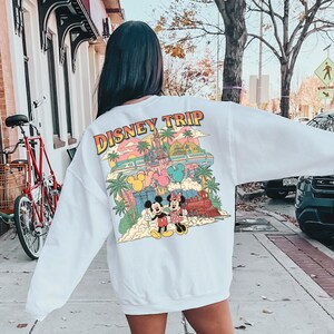 May include: White sweatshirt with a colorful Disney Trip graphic on the back. The graphic features Mickey and Minnie Mouse, a castle, a train, and palm trees. The text "DISNEY TRIP" is in a retro font.