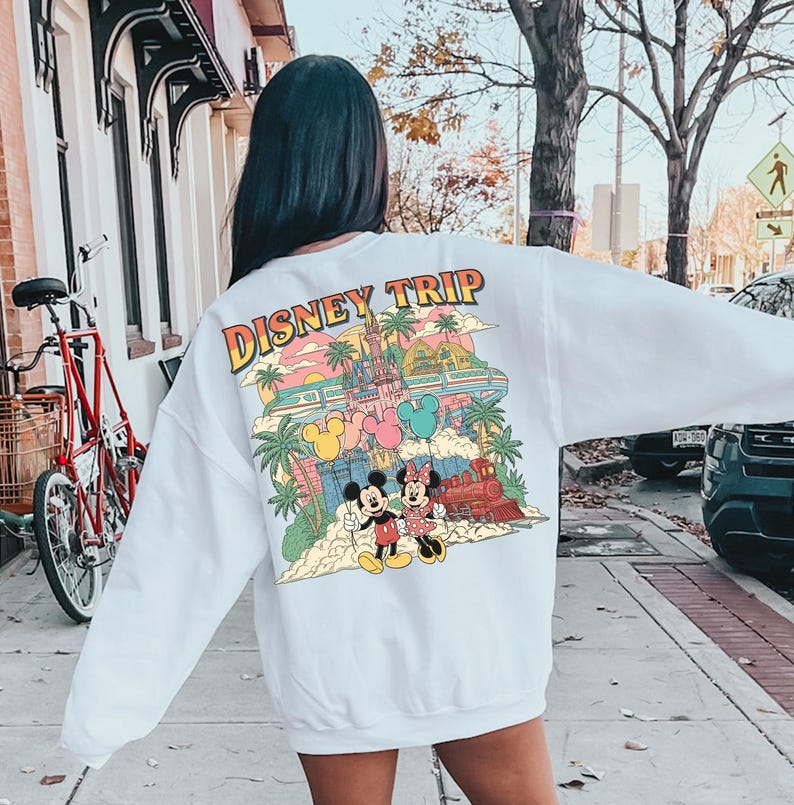 May include: White sweatshirt with a colorful Disney Trip graphic on the back. The graphic features Mickey and Minnie Mouse, a castle, a train, and palm trees. The text "DISNEY TRIP" is in a retro font.
