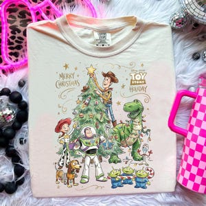 May include: Cream-colored t-shirt featuring a festive Toy Story holiday design. The graphic includes Woody, Buzz Lightyear, Rex, and other characters decorating a Christmas tree. The text "Merry Christmas" and "Toy Story Holiday" are also visible.