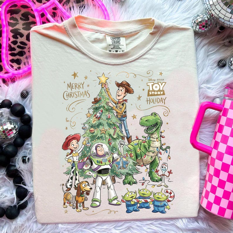 May include: Cream-colored t-shirt featuring a festive Toy Story holiday design. The graphic includes Woody, Buzz Lightyear, Rex, and other characters decorating a Christmas tree. The text "Merry Christmas" and "Toy Story Holiday" are also visible.