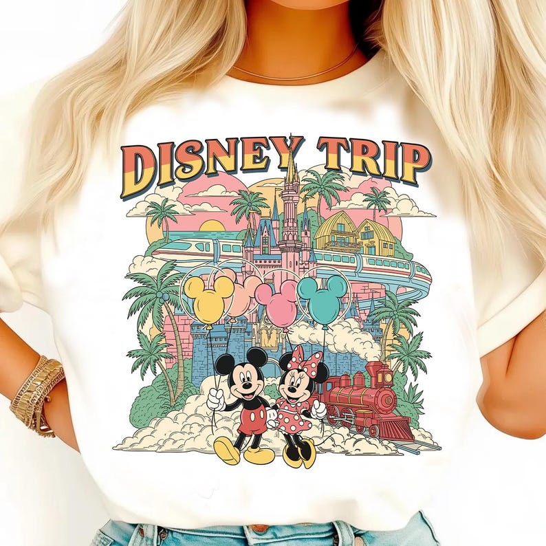 May include: White t-shirt with a colorful graphic that says "DISNEY TRIP". The graphic features Mickey and Minnie Mouse holding balloons, a castle, train, and palm trees. The design has a retro, vintage style.