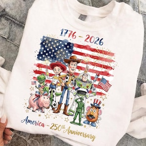 K&ouml;nnte beinhalten: Wei&szlig;es Sweatshirt mit einer Grafik von Zeichentrickfiguren vor einer stilisierten amerikanischen Flagge mit Feuerwerk. Der Text lautet "1776-2026" und "America - 250th Anniversary."