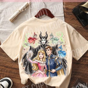 May include: A beige t-shirt with a colourful illustration of fairy tale characters. The design shows a princess, a prince, and a villain taking a selfie, with the text "ORIGINAL DESIGNER CALIGRAPHICDESIGN" at the top.