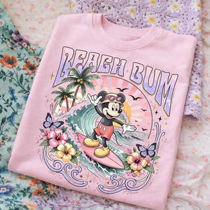 May include: Pink sweatshirt with a graphic of Mickey Mouse surfing a wave. The design includes palm trees, butterflies, and the words "Beach Bum". The sweatshirt has a crew neck and long sleeves.