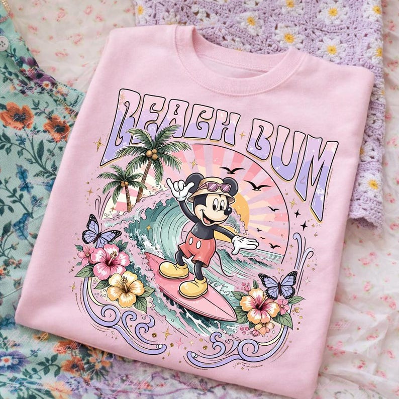 May include: Pink sweatshirt with a graphic of Mickey Mouse surfing a wave. The design includes palm trees, butterflies, and the words "Beach Bum". The sweatshirt has a crew neck and long sleeves.