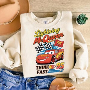 May include: Cream-colored sweatshirt featuring a graphic of Lightning McQueen from the movie Cars. The design includes the text "Lightning McQueen", "95", and "Think Fast". The red race car is flanked by checkered flags.