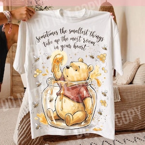 May include: White t-shirt featuring a cartoon bear in a honey jar, surrounded by bees and honeycombs. The shirt has the text "Sometimes the smallest things take up the most room in your heart."