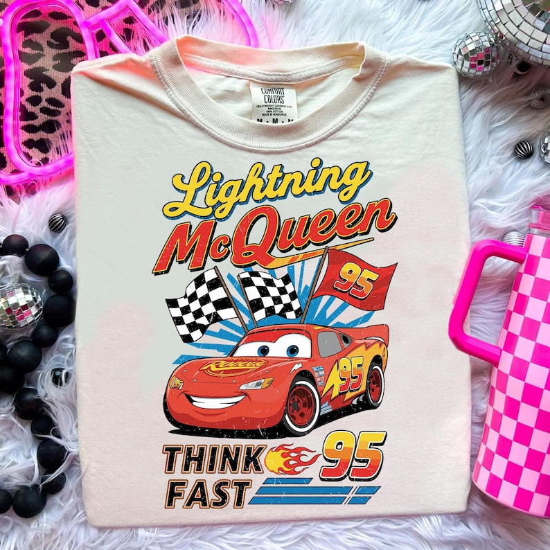 May include: Cream-colored t-shirt featuring a graphic of Lightning McQueen from the Cars movie. The design includes the text "Lightning McQueen", "95", and "Think Fast". The red race car is depicted with a smiling face and checkered flags.