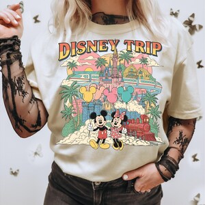 May include: Cream-colored t-shirt with a colorful graphic design featuring Mickey and Minnie Mouse, a castle, train, and the text "DISNEY TRIP". The design includes palm trees, balloons, and a sunset.