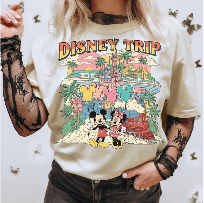 May include: Cream-colored t-shirt with a colorful graphic design featuring Mickey and Minnie Mouse, a castle, train, and the text "DISNEY TRIP". The design includes palm trees, balloons, and a sunset.