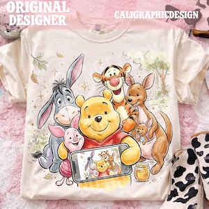 May include: Cream-colored t-shirt featuring a colorful illustration of Winnie the Pooh and friends, including Piglet, Eeyore, Tigger, and Roo. The characters are holding a phone displaying a group photo. The text "ORIGINAL DESIGNER" and "CALIGRAPHICDESIGN" are visible.
