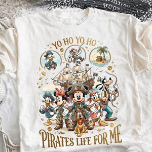 Peut inclure: T-shirt blanc avec un motif coloré sur le thème des pirates. Le design comprend Mickey Mouse, Minnie Mouse, Donald Duck et Dingo déguisés en pirates, avec les textes "YO HO YO HO" et "PIRATES LIFE FOR ME".