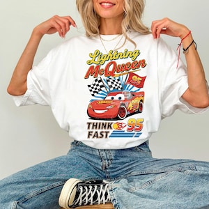May include: White t-shirt featuring Lightning McQueen from the Cars movie. The design includes the text "Lightning McQueen" in yellow and red, a red race car, checkered flags, and the text "THINK FAST 95".