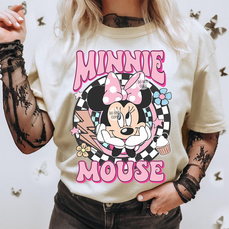 May include: Cream-colored t-shirt featuring a graphic of Minnie Mouse with a pink bow and the text "MINNIE MOUSE" in pink bubble letters. The design includes a checkered background, lightning bolt, flowers, and a cupcake.