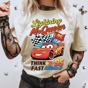 May include: Cream-colored t-shirt featuring a graphic of Lightning McQueen from the Cars movie. The design includes the text "Lightning McQueen" and "Think Fast 95" with checkered flags and a red race car.