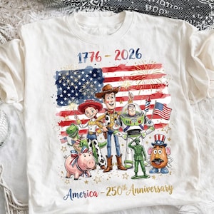 K&ouml;nnte beinhalten: Wei&szlig;es T-Shirt mit einer farbenfrohen Grafik der amerikanischen Flagge, Feuerwerk und Zeichentrickfiguren. Der Text "1776-2026" und "America - 250th Anniversary" sind ebenfalls auf dem T-Shirt abgebildet.