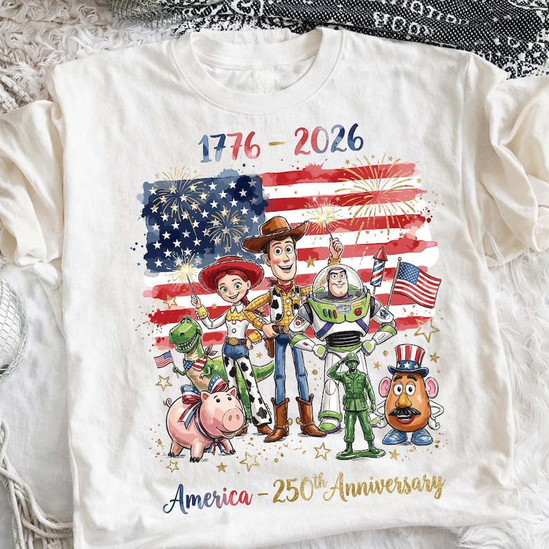 K&ouml;nnte beinhalten: Wei&szlig;es T-Shirt mit einer farbenfrohen Grafik der amerikanischen Flagge, Feuerwerk und Zeichentrickfiguren. Der Text "1776-2026" und "America - 250th Anniversary" sind ebenfalls auf dem T-Shirt abgebildet.
