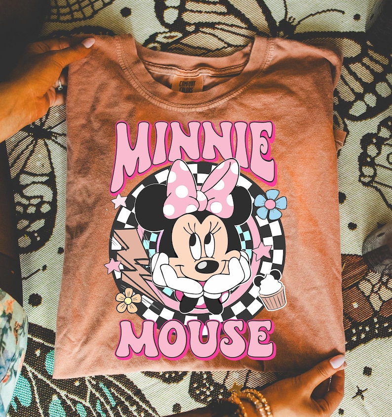 May include: A peach-colored t-shirt featuring a graphic of Minnie Mouse with a pink polka-dot bow, surrounded by flowers, stars, and a lightning bolt. The text "MINNIE MOUSE" is in pink, retro-style lettering.
