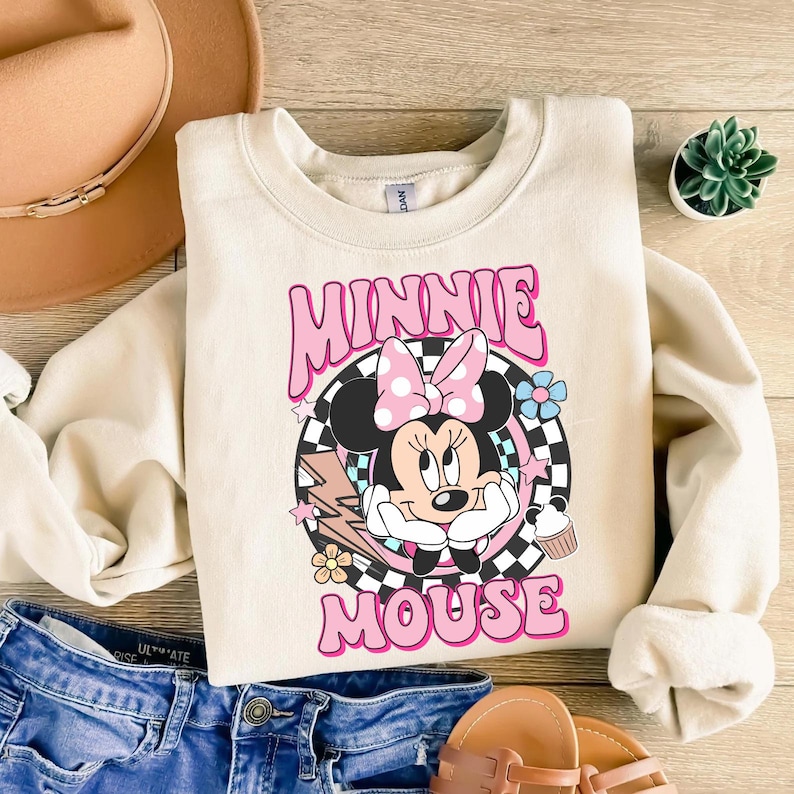 May include: Cream-colored sweatshirt featuring a graphic of Minnie Mouse with a pink bow and the words "MINNIE MOUSE" in pink. The design includes a checkered background, flowers, and a cupcake. The sweatshirt has long sleeves.