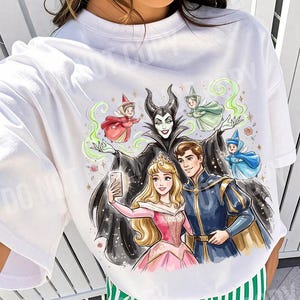 May include: White crew neck t-shirt with a colourful illustration of Disney characters taking a selfie. The design includes Maleficent, Aurora, Prince Phillip, and fairies. Text at the top reads "ORIGINAL DESIGNER CALIGRAPHICDESIGN".