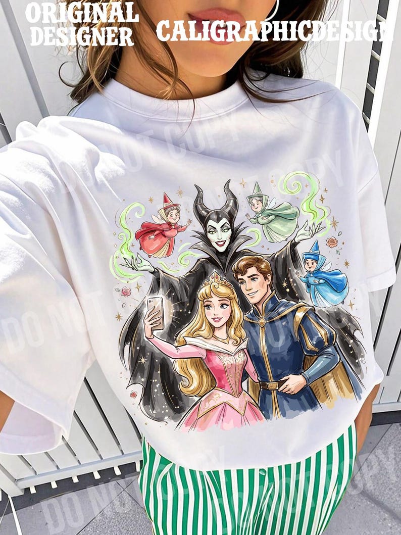 May include: White crew neck t-shirt with a colourful illustration of Disney characters taking a selfie. The design includes Maleficent, Aurora, Prince Phillip, and fairies. Text at the top reads "ORIGINAL DESIGNER CALIGRAPHICDESIGN".