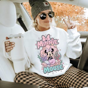 May include: White sweatshirt featuring a Minnie Mouse mermaid design in a seashell. The graphic includes the words "Minnie Mouse" in pink and blue. The sweatshirt is worn with a gray beanie, sunglasses, and plaid pants.