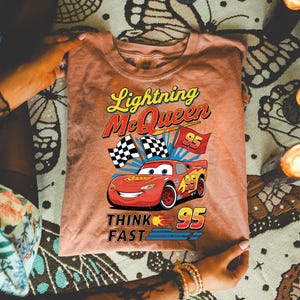 May include: A peach-colored t-shirt featuring Lightning McQueen from the movie Cars. The graphic includes the text "Lightning McQueen", "95", and "Think Fast", along with a smiling red race car and checkered flags.