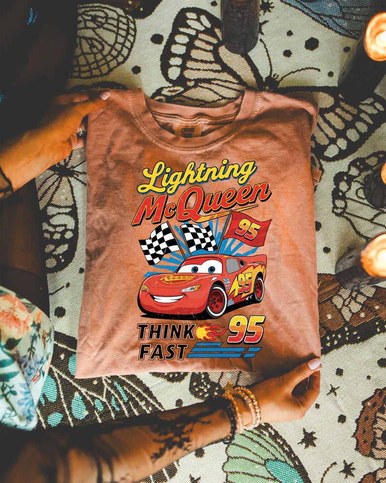 May include: A peach-colored t-shirt featuring Lightning McQueen from the movie Cars. The graphic includes the text "Lightning McQueen", "95", and "Think Fast", along with a smiling red race car and checkered flags.