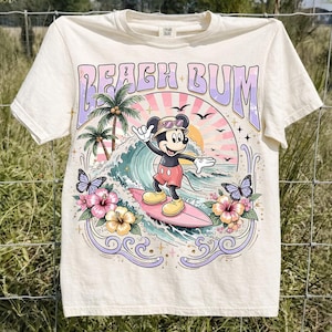 May include: Cream-colored t-shirt featuring a colorful graphic of Mickey Mouse surfing. The design includes the words "Beach Bum", palm trees, waves, butterflies, and flowers. The shirt has short sleeves and a crew neck.